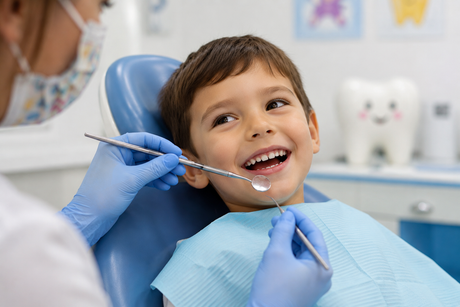 Un bambino sorride sulla poltrona del dentista mentre quest'ultimo gli esamina i denti con uno specchietto e le mani guantate.