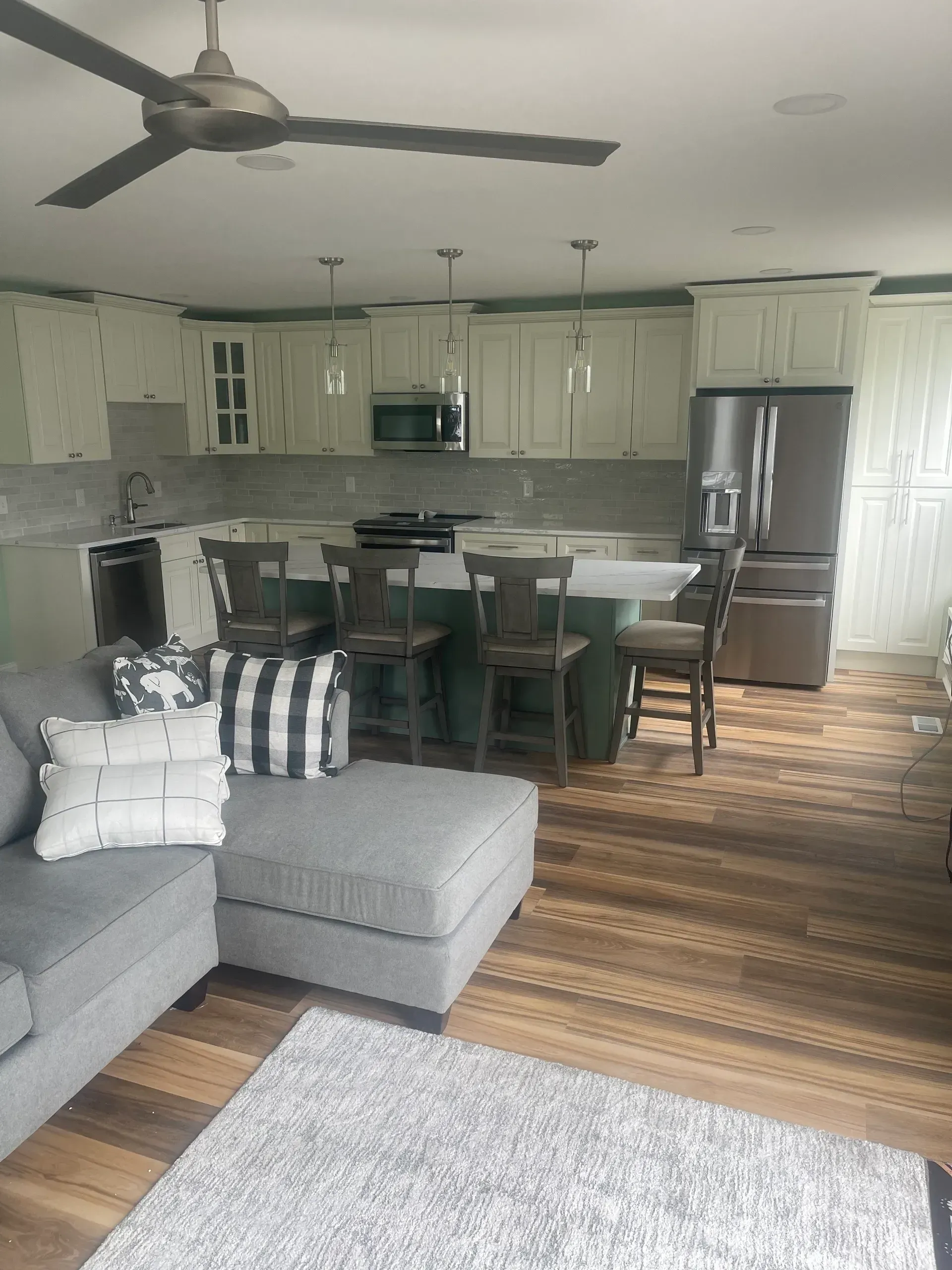 Kitchen with white cabinets, green island, and stainless steel appliances. Gray sectional sofa and wooden floors.