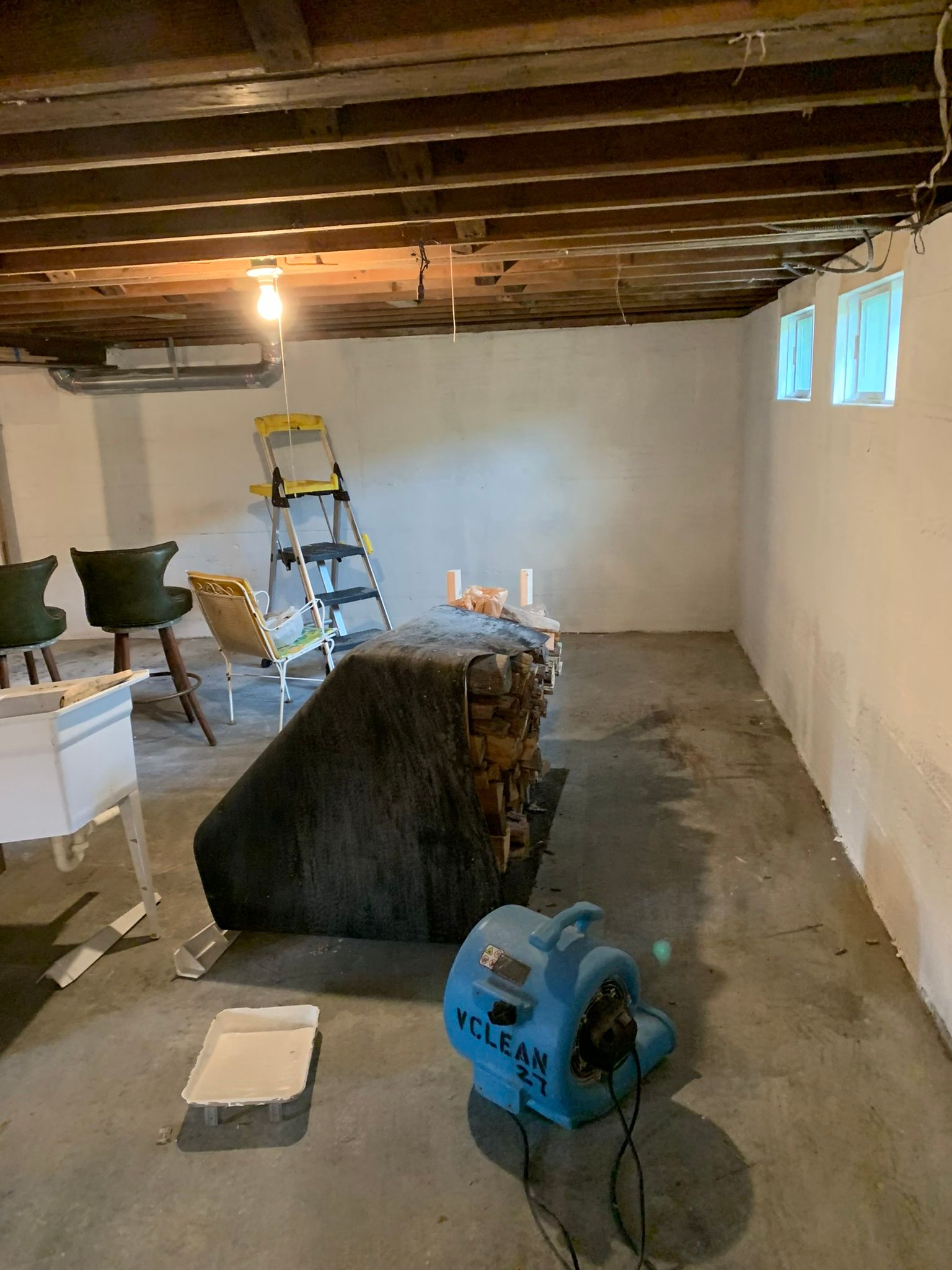 Unfinished basement with exposed ceiling, ladder, tools, and furniture being moved or covered for renovation.