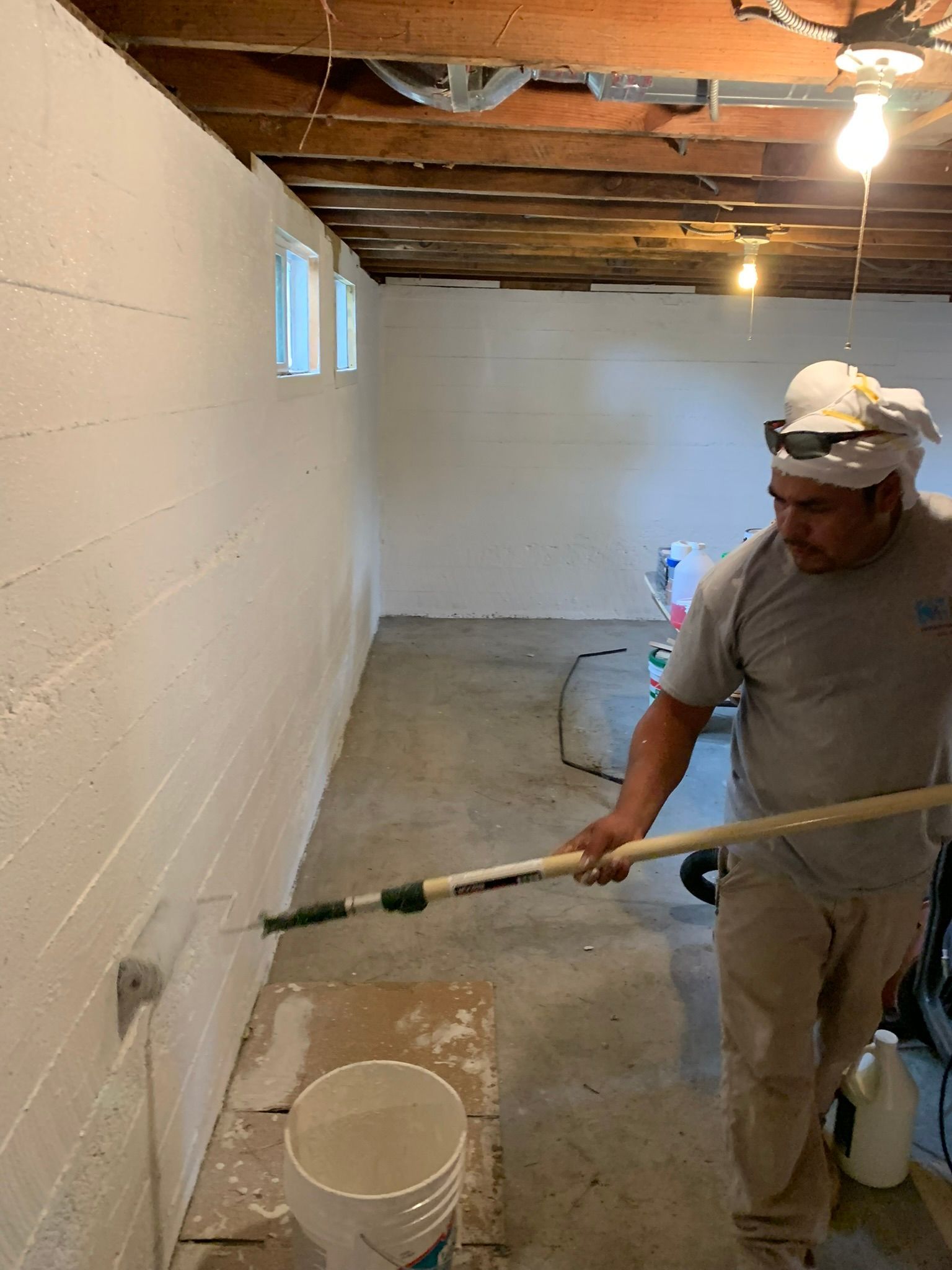 Worker painting a basement wall with a roller near a bucket and drop cloth on the floor