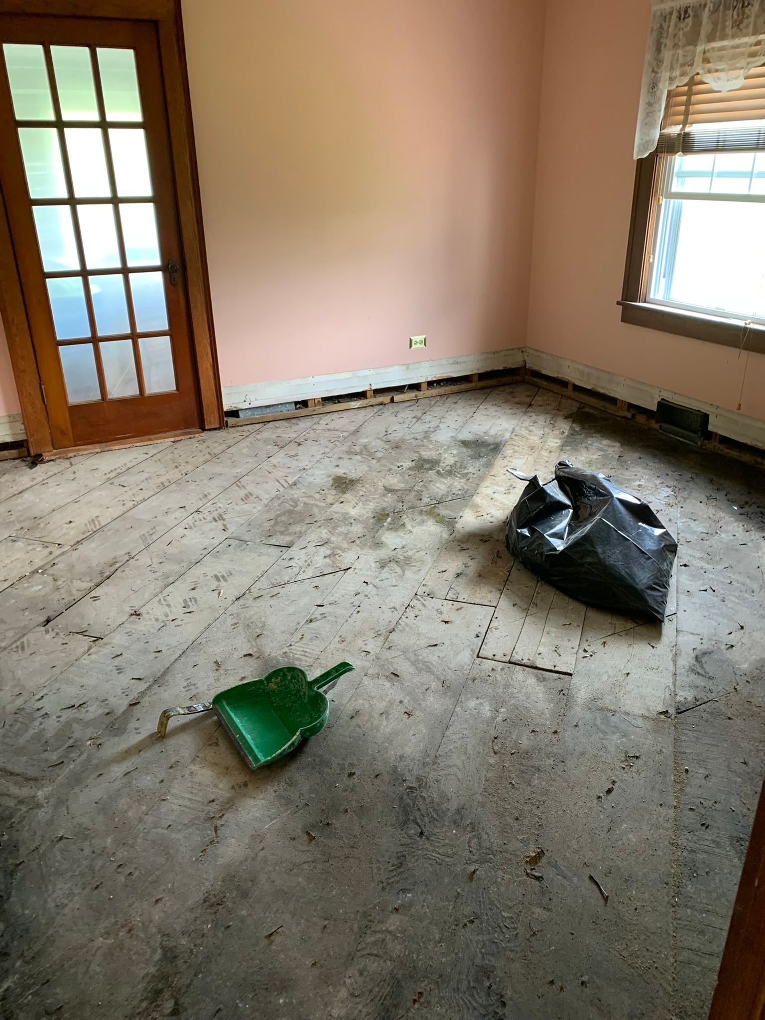 Empty room with dusty floor, a green dustpan, and a black trash bag near the wall.
