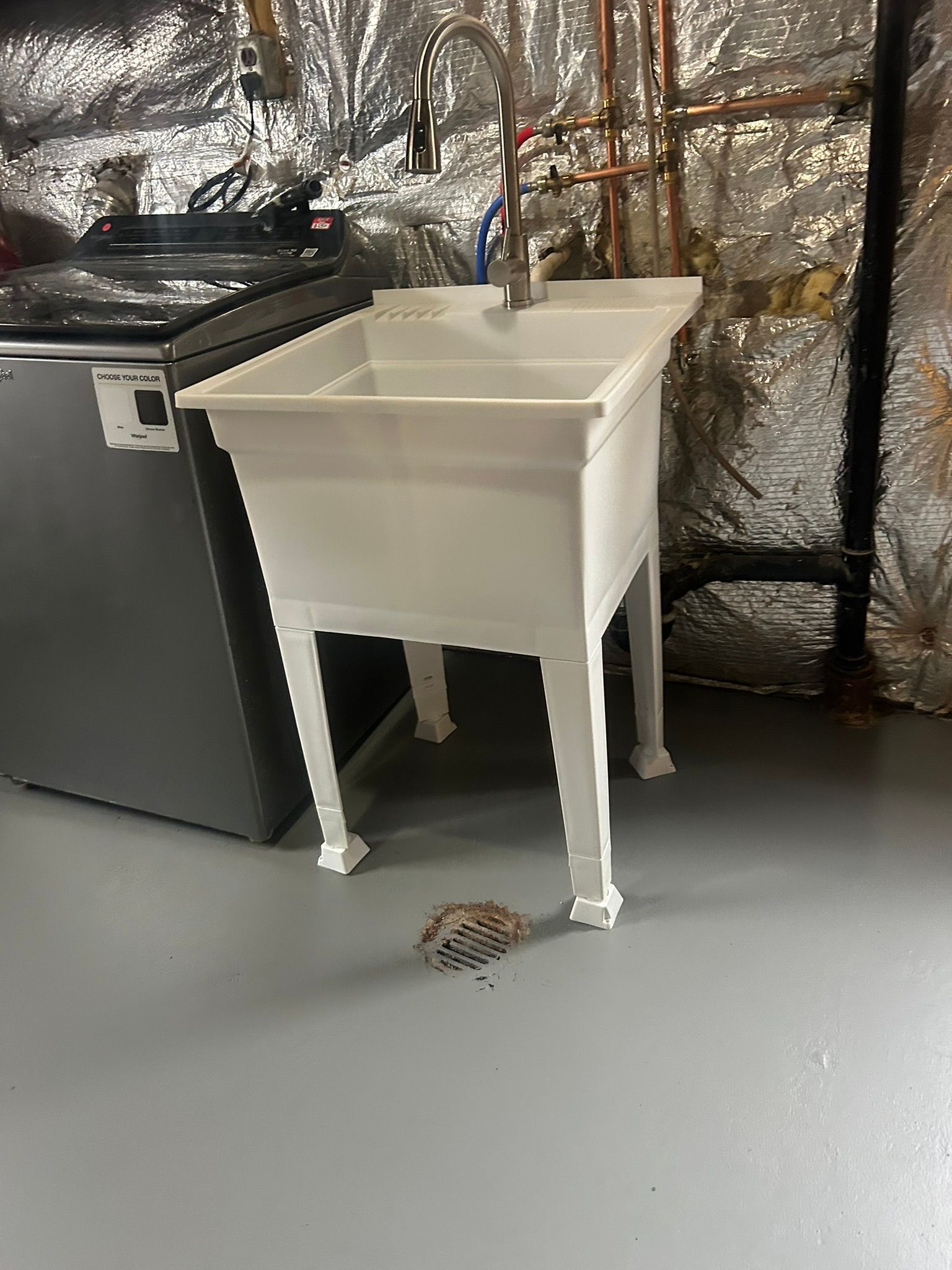 White utility sink in a basement beside a black appliance, with foil-covered insulated walls behind it.