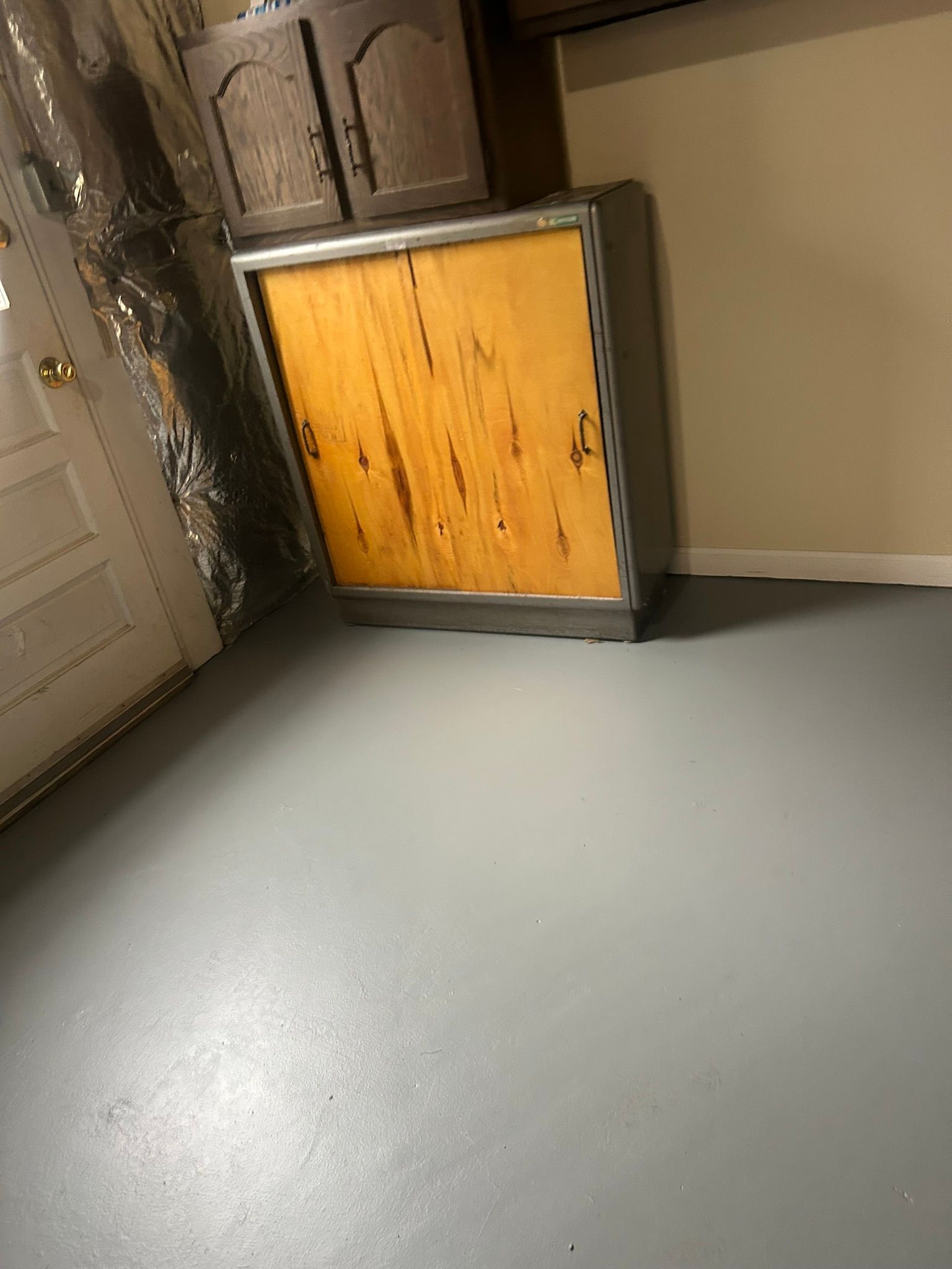 Empty room corner with a gray floor, beige walls, dark cabinets, and a small wooden cabinet or heater unit.