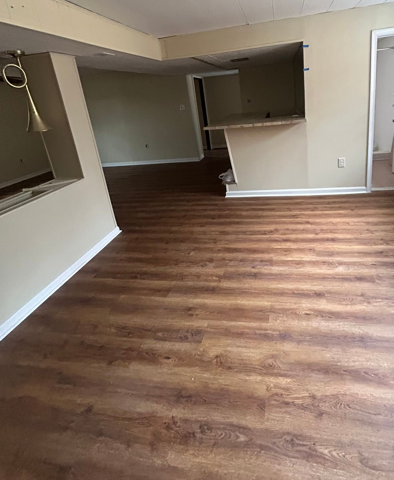Empty living room with brown wood flooring, beige walls, and a kitchen pass-through.