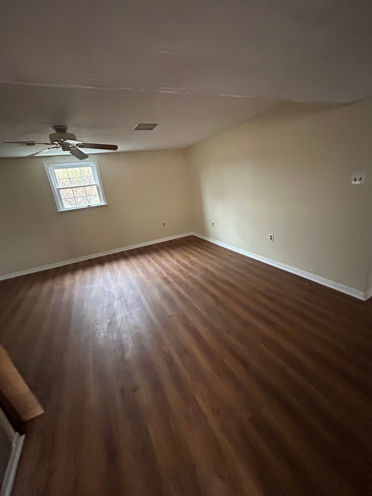Empty beige room with wood floors, ceiling fan, and a small window on the far wall.