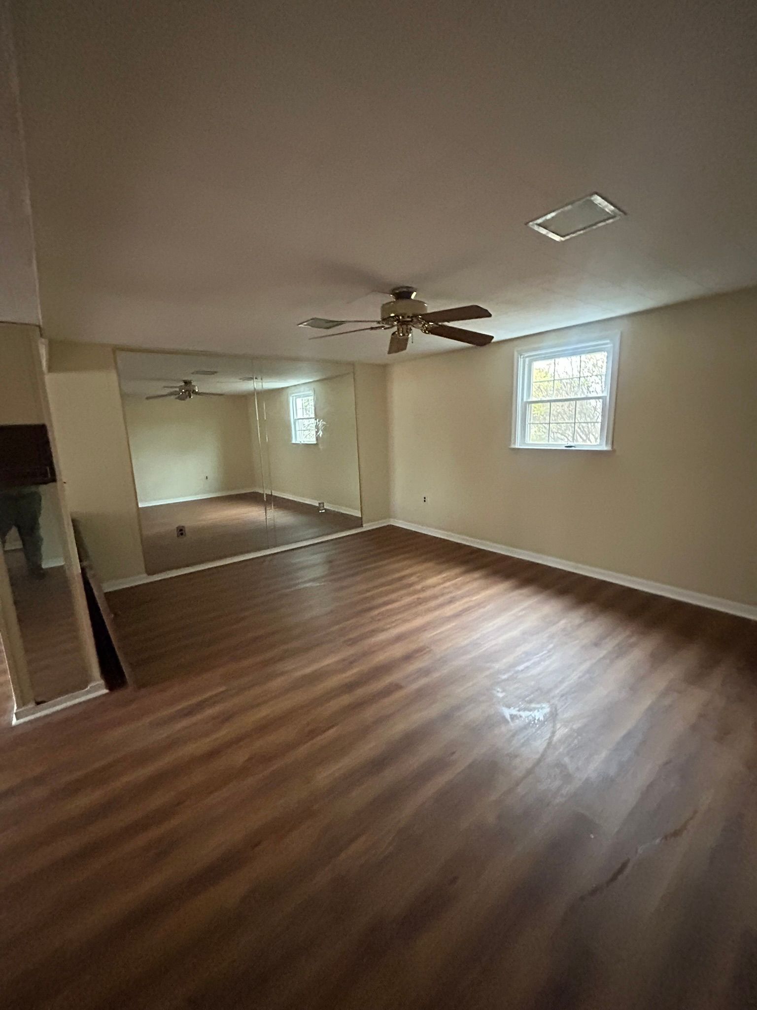 Empty living room with wood floors, ceiling fan, mirrored wall, and a small window.