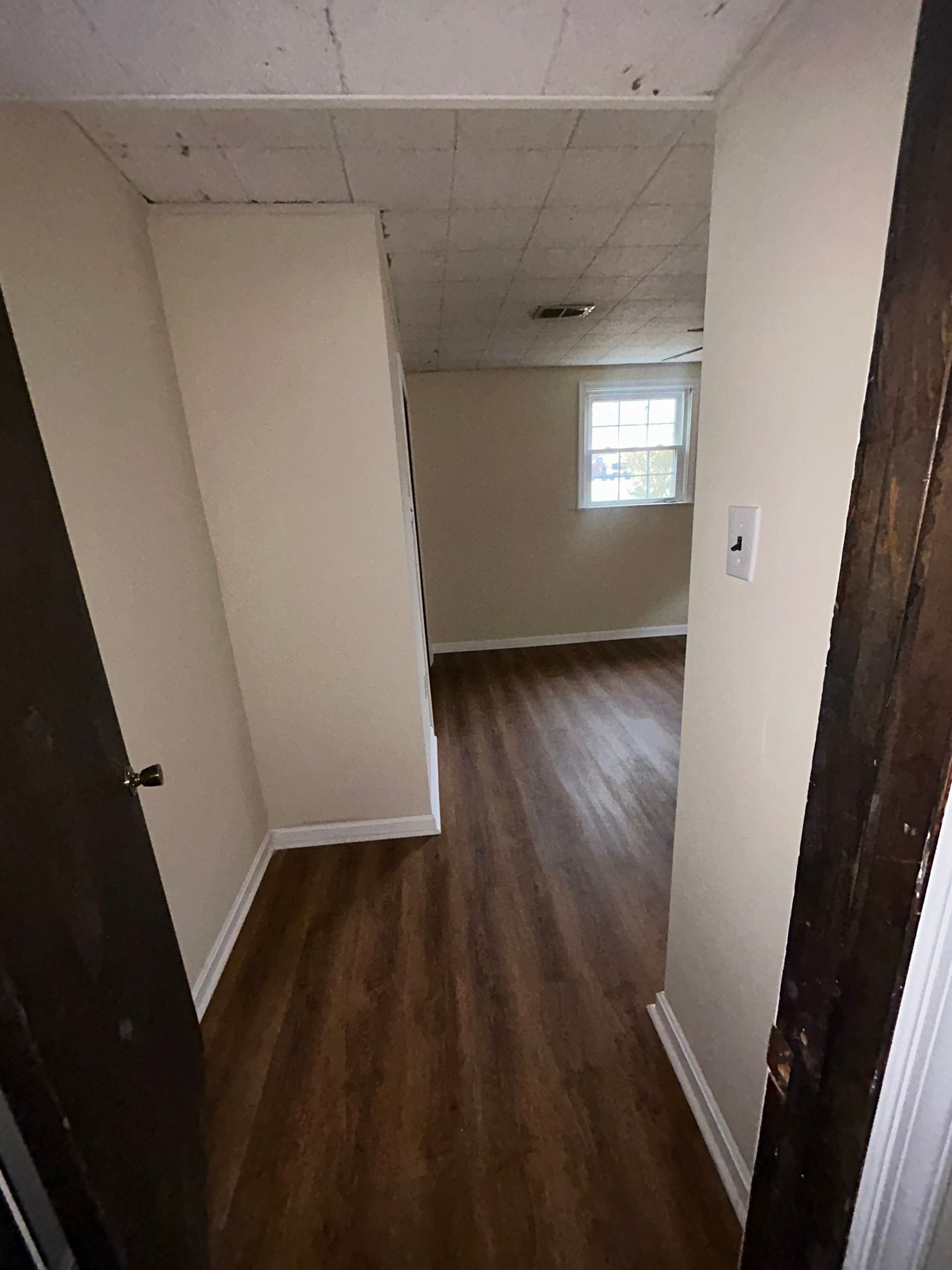 Narrow hallway with dark wood floors, white walls, and a small window at the end.