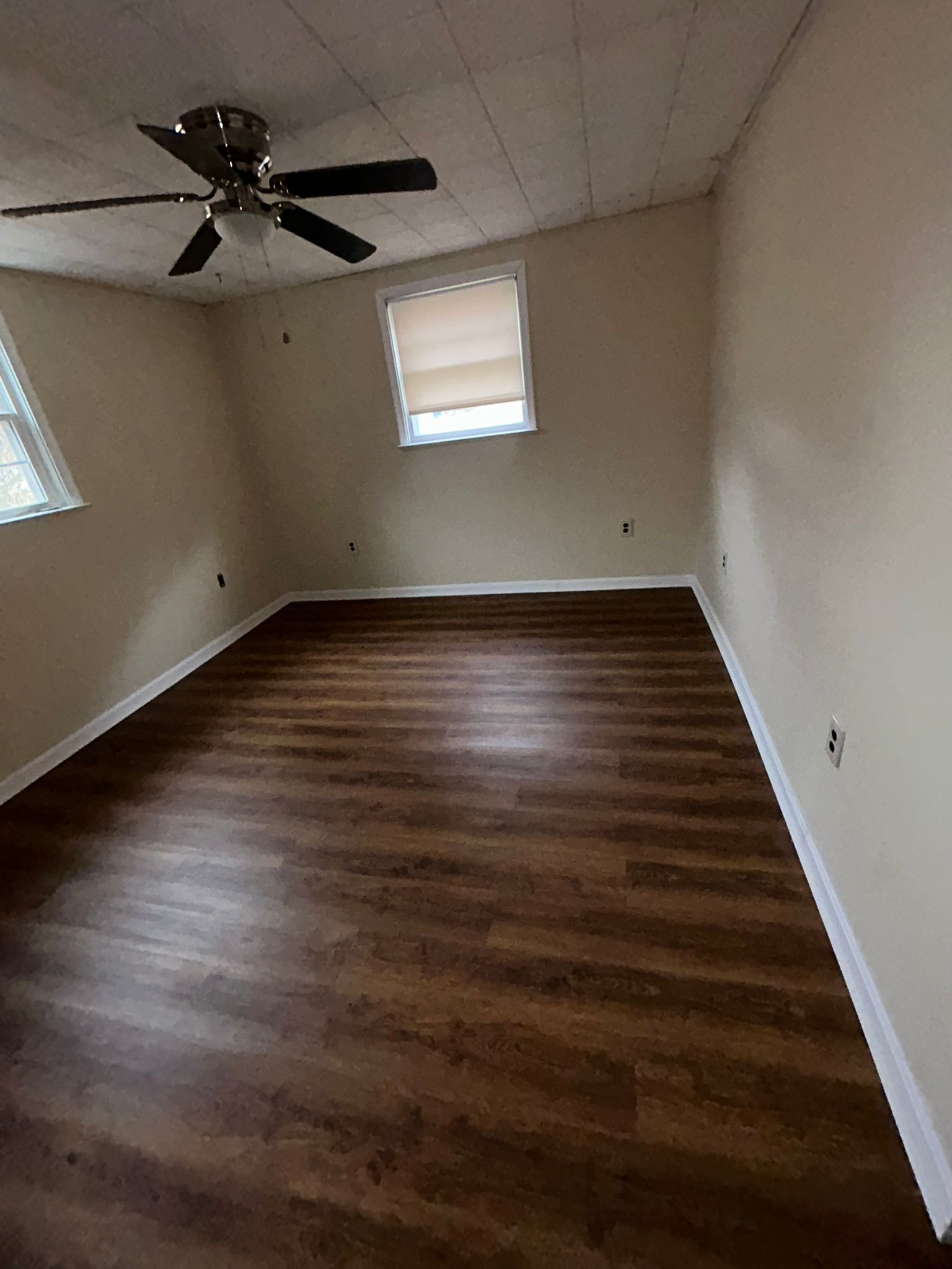 Empty room with brown wood flooring, beige walls, ceiling fan, and two windows.