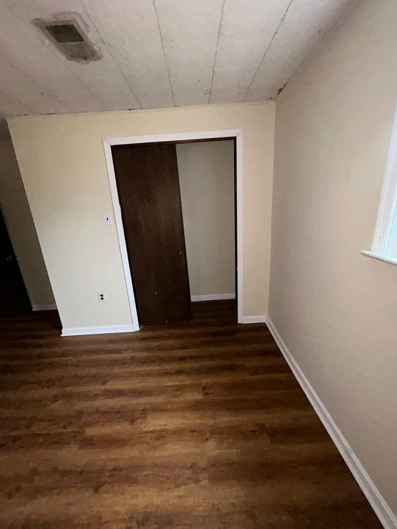 Empty room with wood floors, beige walls, open closet, and a window on the right.