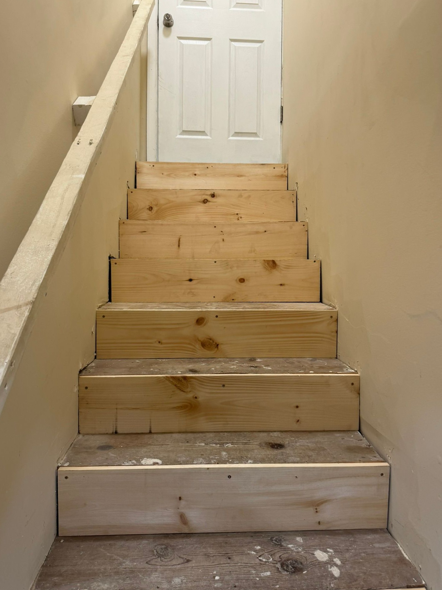 Narrow unfinished wooden staircase leading up to a white door in a beige hallway.