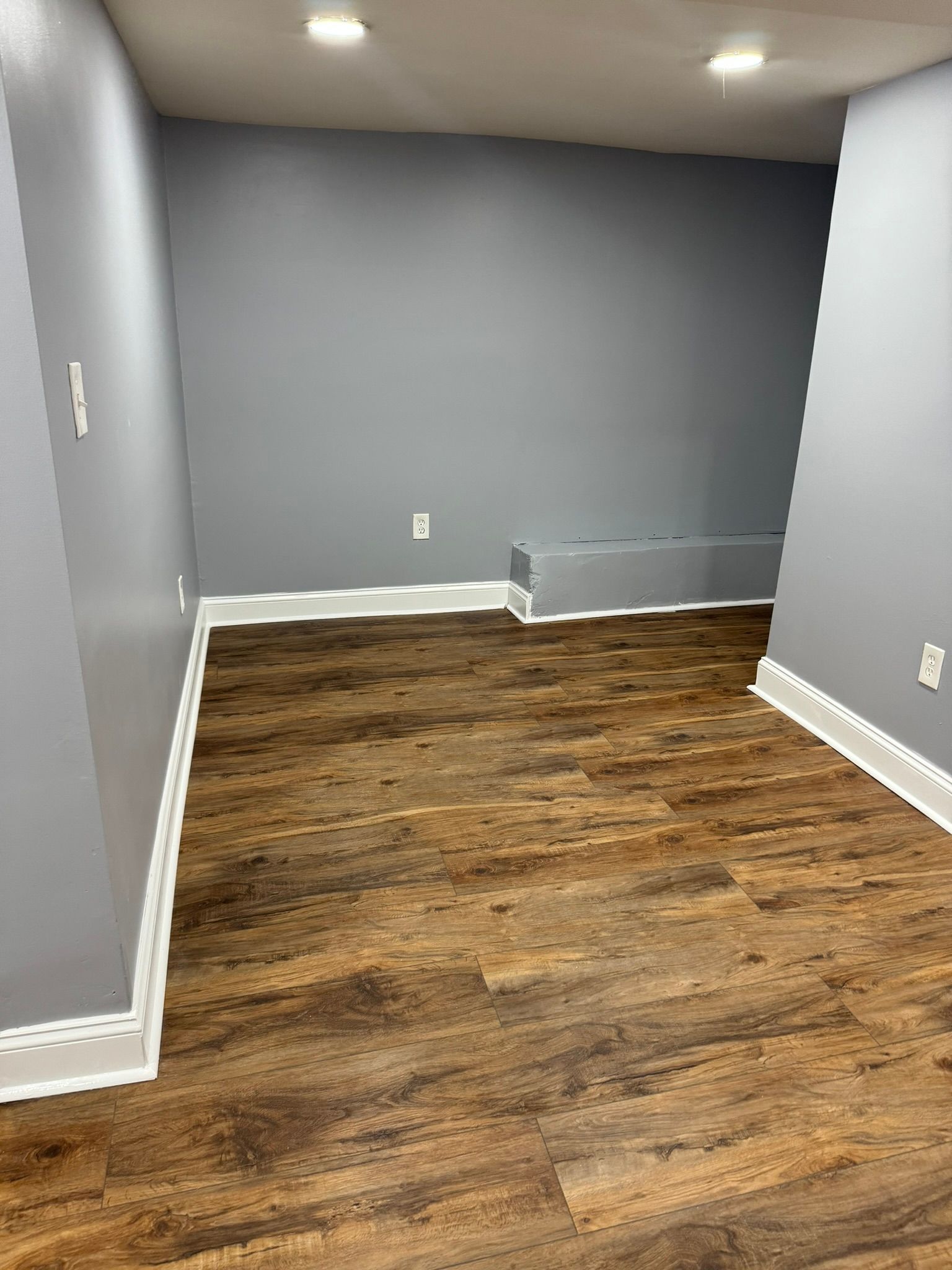 Empty room with gray walls, wood-look floor, white trim, and recessed ceiling lights.