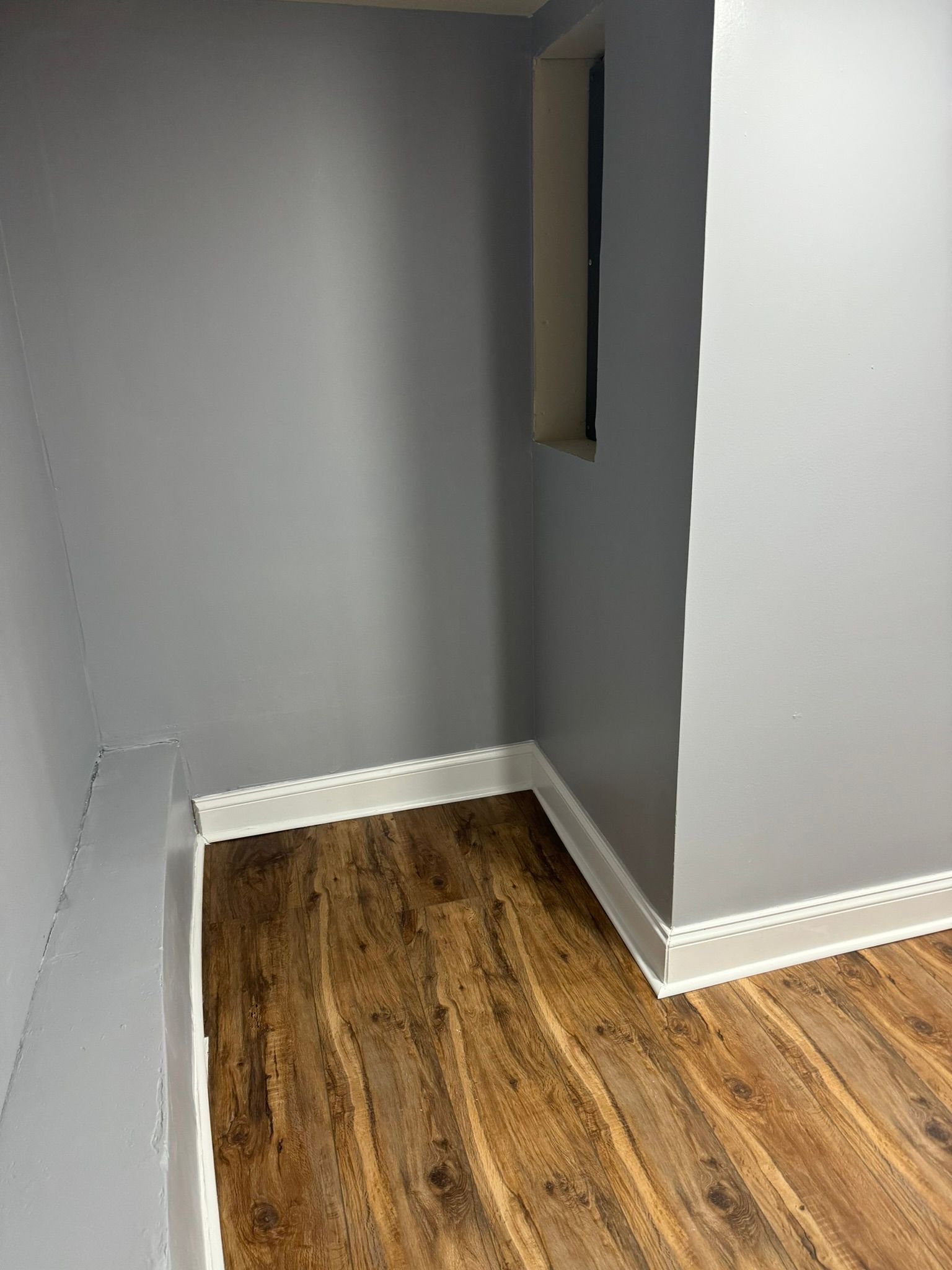 Empty gray corner room with wood floor and white baseboards, plus a narrow wall opening.