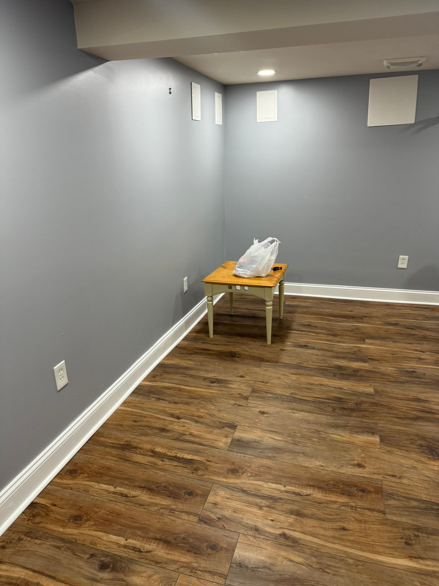 Empty gray room with wood flooring, white baseboards, small windows, and a small table with items near the back wall