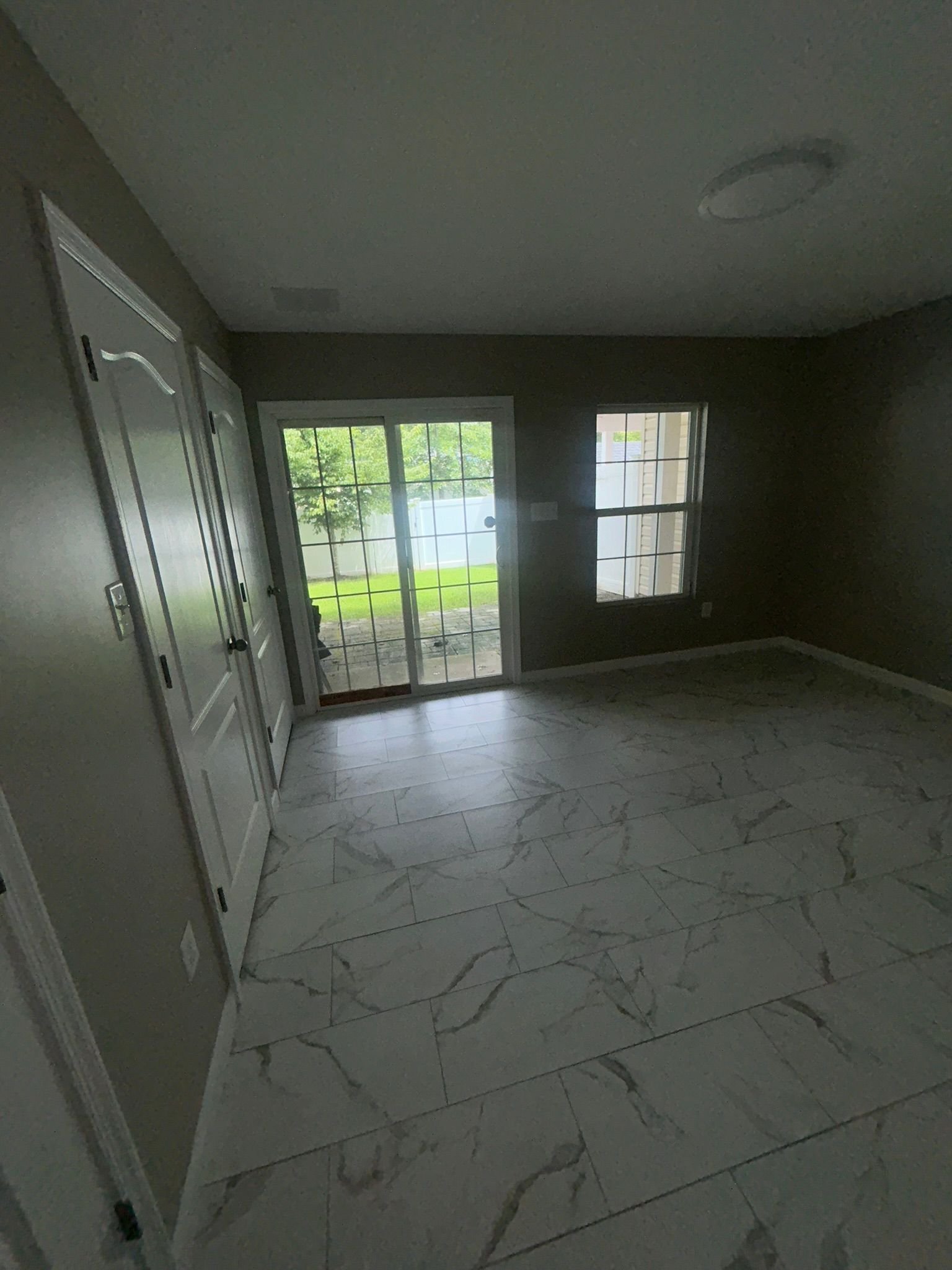 Empty living room with tile floor, gray walls, and glass doors opening to a green backyard.