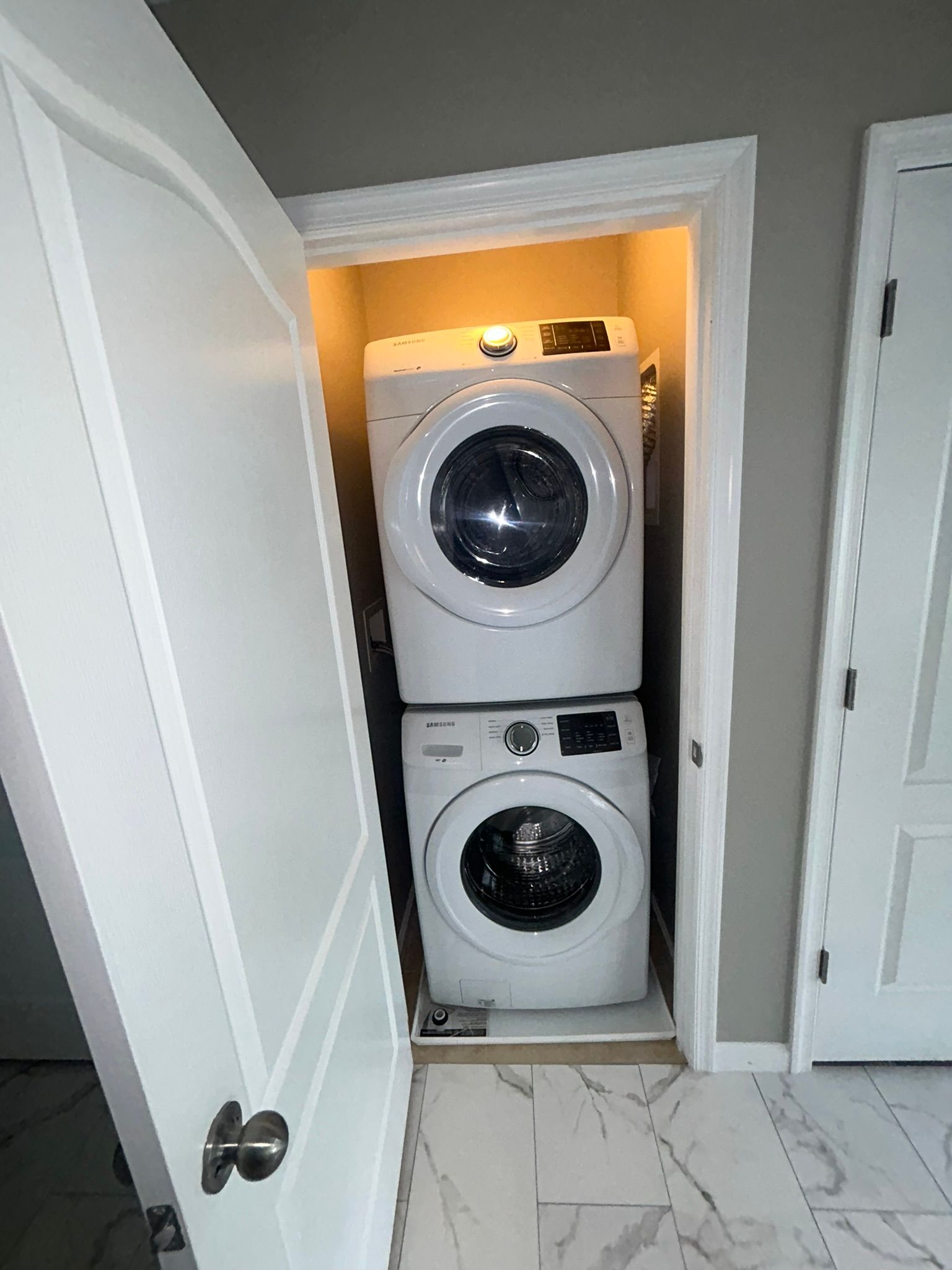 Stacked washer and dryer in a narrow closet, with white doors and a lit alcove.