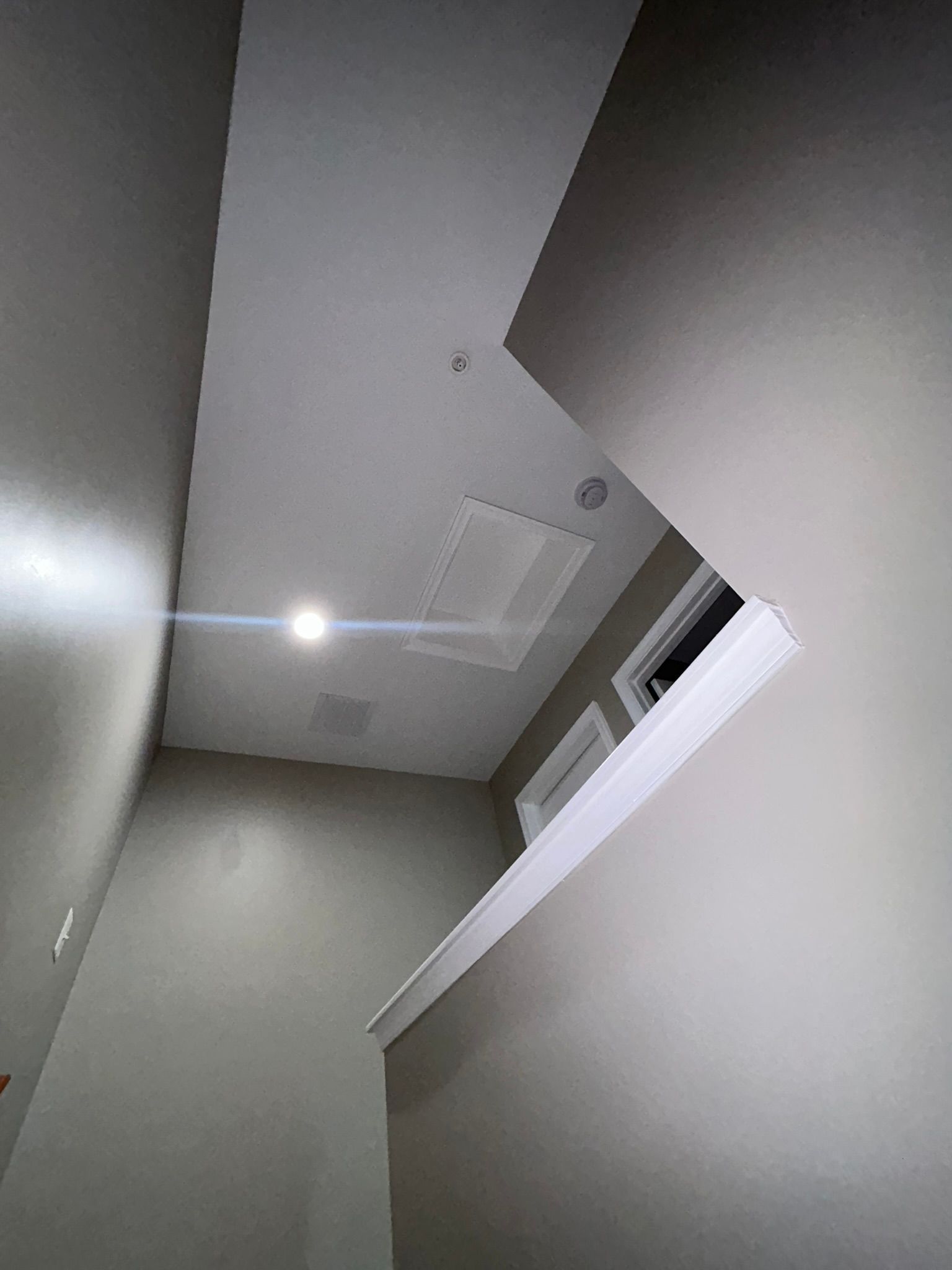 Upward view of a narrow stairwell with gray walls, white ceiling, vents, and a bright light fixture