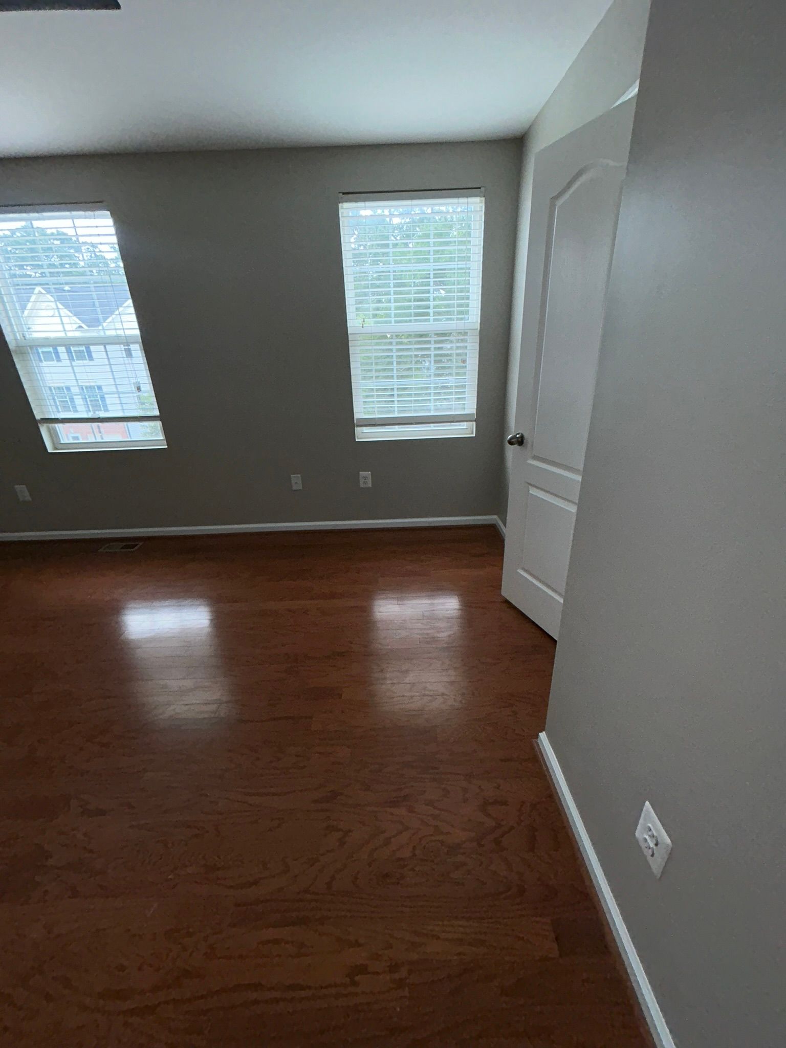 Empty room with hardwood floors, gray walls, two windows, and a white door.