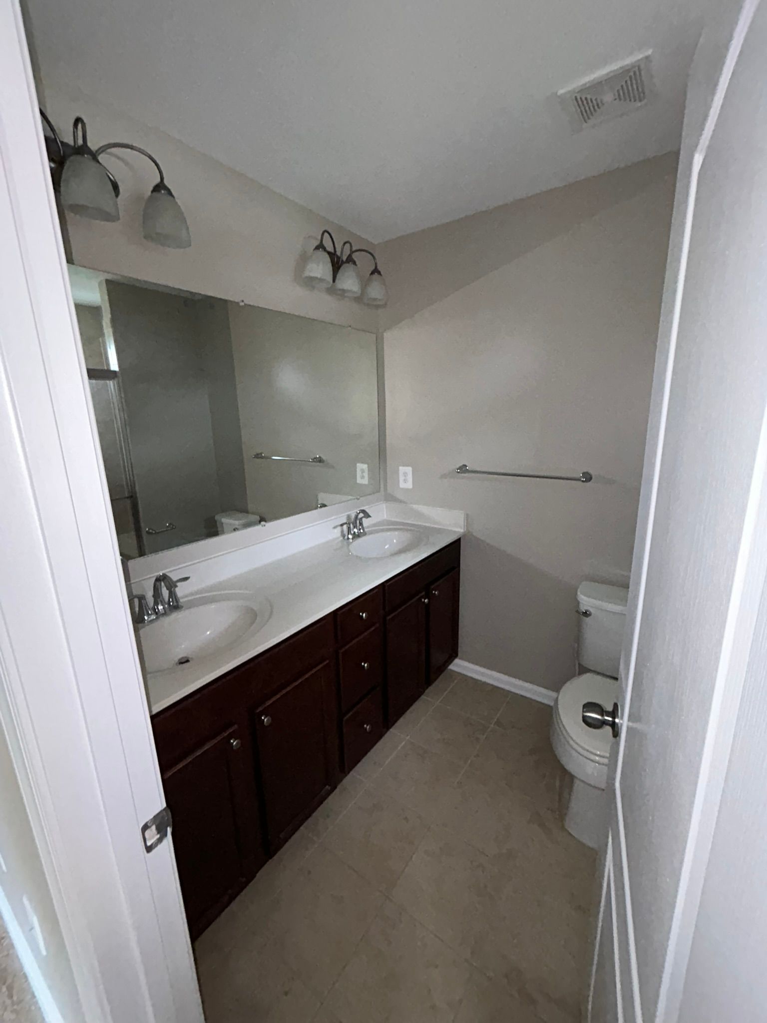 Small bathroom with dark wood vanity, large mirror, white sink, toilet, and beige tile floor.