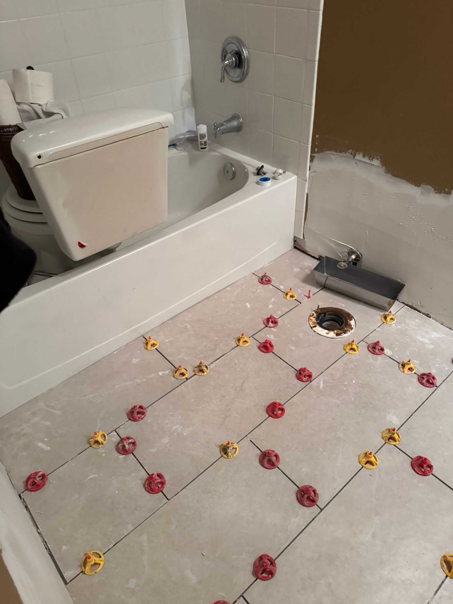Bathroom floor under repair with white square tiles and scattered pink and yellow flowers around a drain.