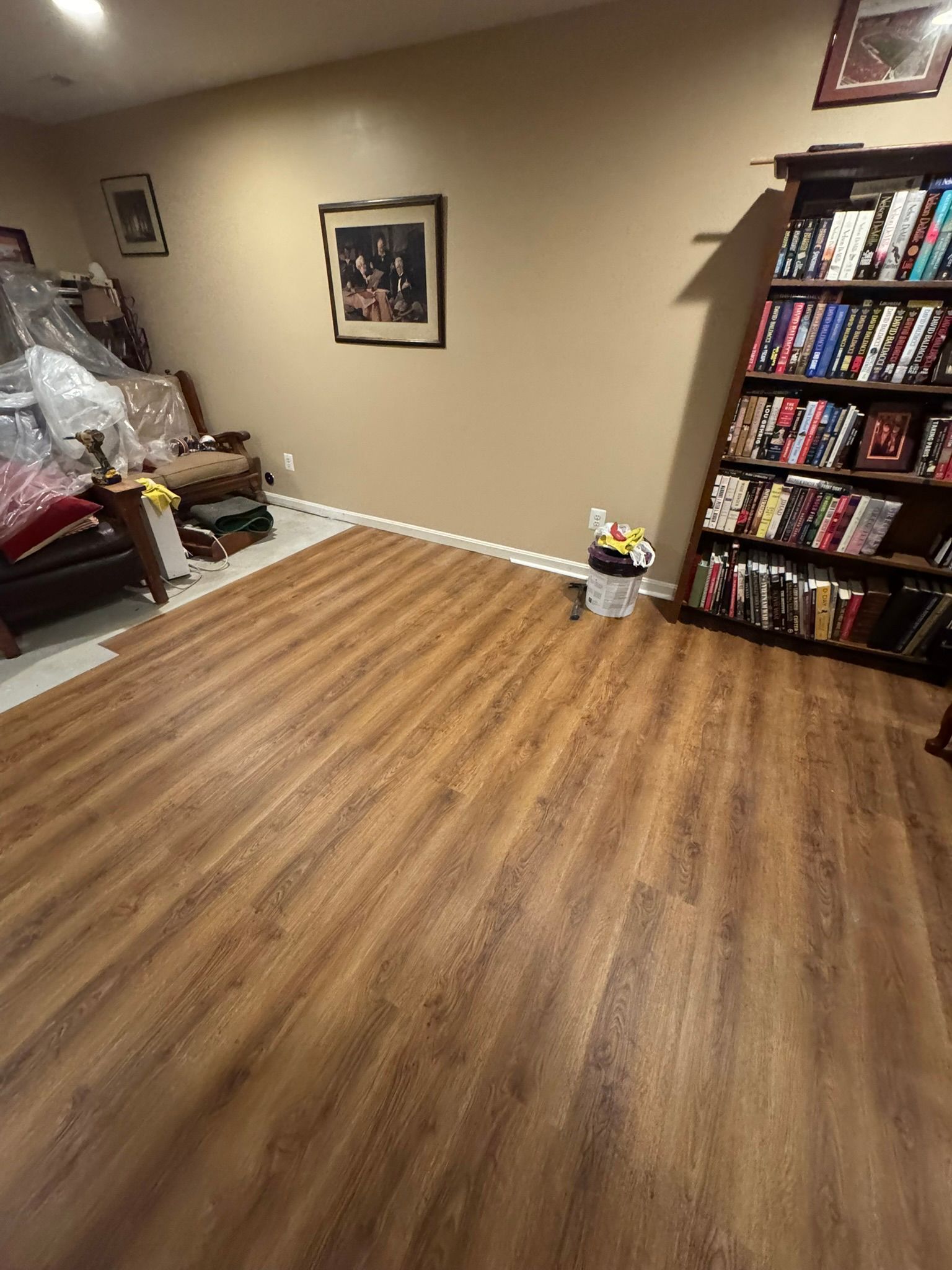 Empty room with wood-pattern floor, beige walls, framed art, and a bookshelf on the right.