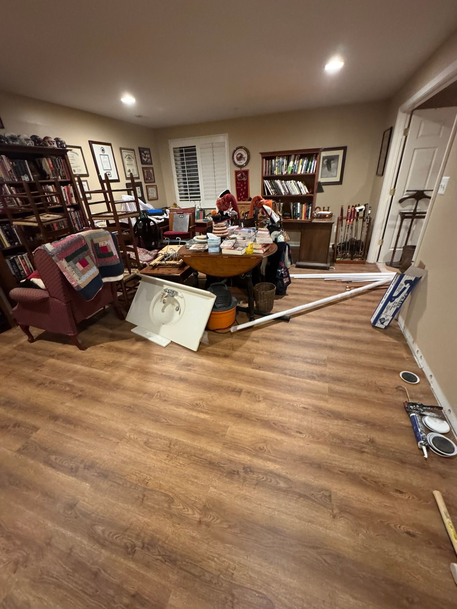 Cluttered living room with furniture, a table of items, and wood flooring under warm ceiling lights