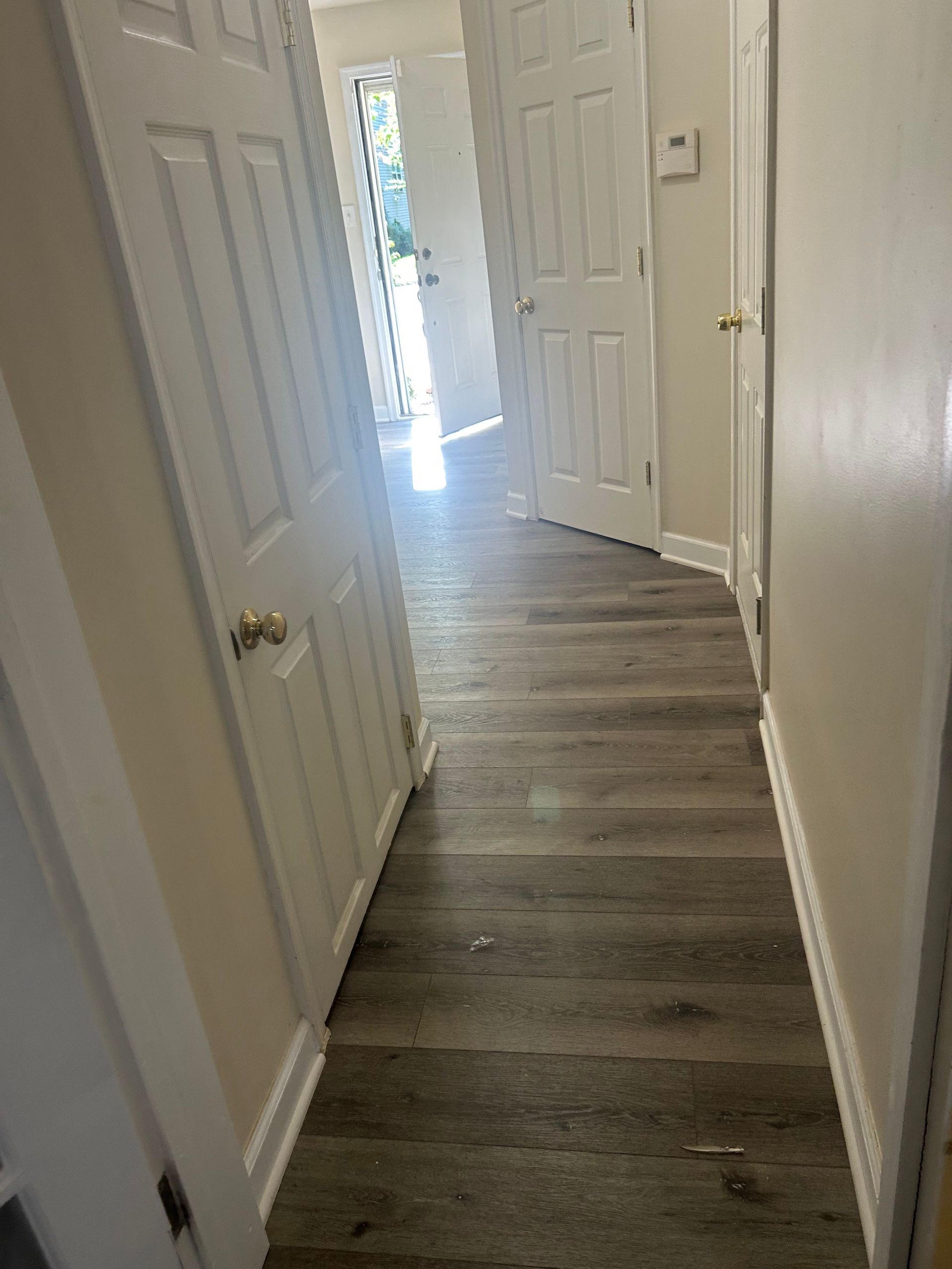 Narrow hallway with white doors and gray wood flooring leading to a bright front doorway
