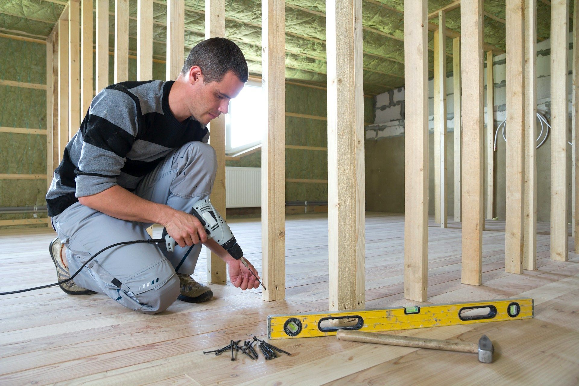 Worker kneeling with drill on a wooden floor under construction, beside a yellow level and exposed wall studs