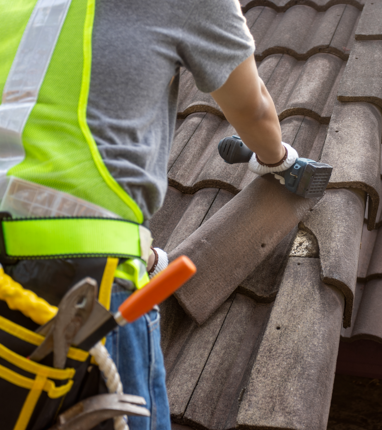 Worker installing roof tiles on a tiled roof, wearing a safety vest and tool belt.