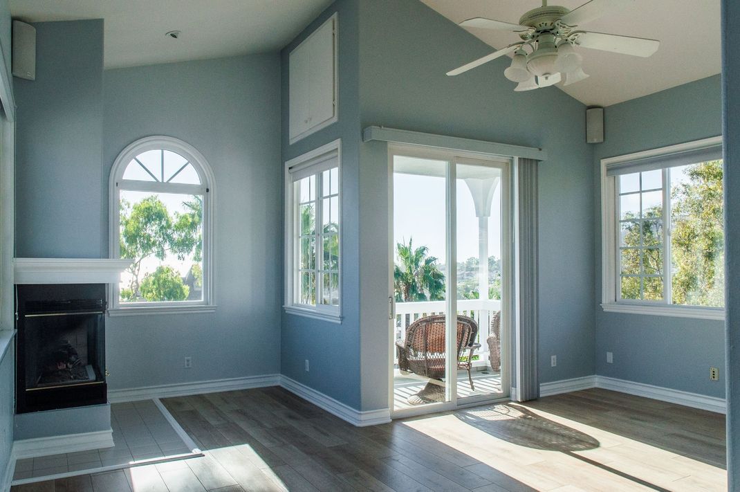 Bright empty living room with gray walls, white trim, fireplace, large windows, and ceiling fan.