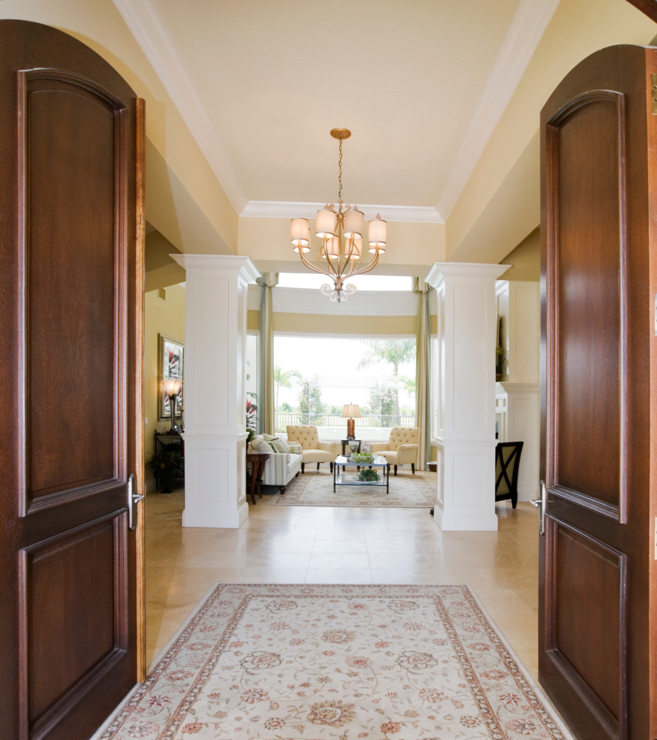 Elegant foyer with open double doors, chandelier, rug, and bright living area beyond.
