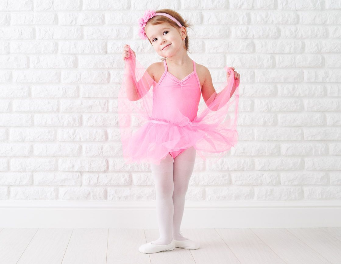 A little girl in a pink tutu and gloves is standing in front of a white brick wall.