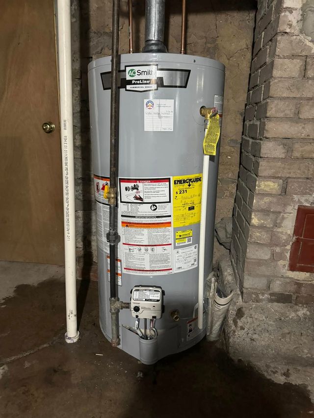 Gray water heater in a basement against a brick wall, with pipes and safety labels.