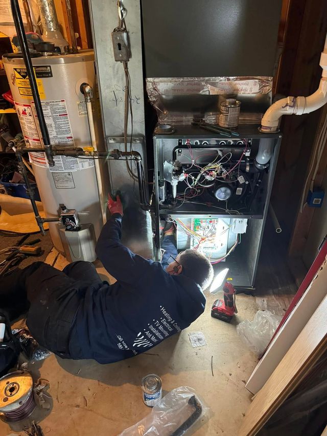 Person kneeling inside a cramped utility space, repairing a furnace with tools and exposed pipes.