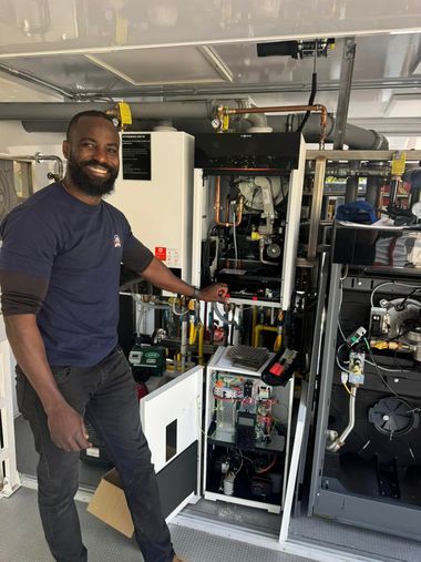 Man standing beside open equipment cabinet in a workshop, holding a tool and smiling.