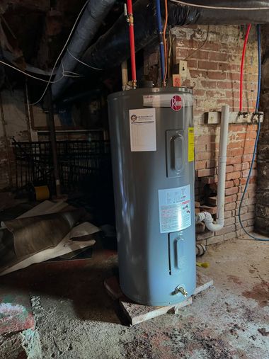 Gray water heater in a basement utility area with exposed pipes and brick wall