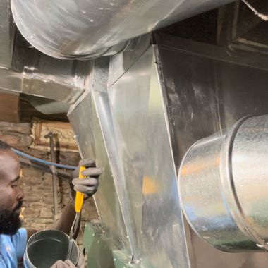 Worker installing shiny metal HVAC ductwork in a ceiling space, using a tool near a large round vent.