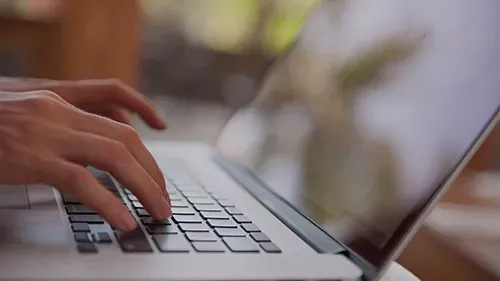Hands typing on a laptop keyboard beside a tilted screen in a soft-focus indoor setting