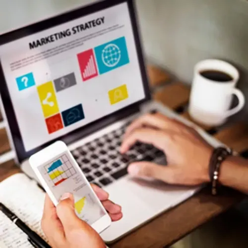 Hand using smartphone beside laptop showing a marketing strategy presentation, with coffee cup on desk