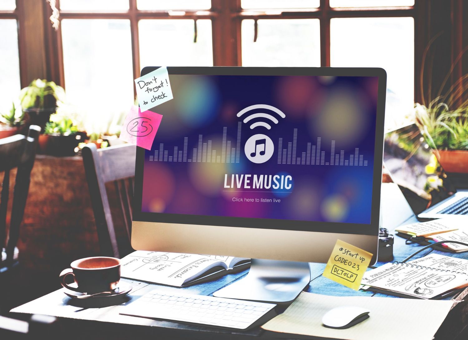 Desk with computer displaying a “Live Music” screen, surrounded by notes, papers, and coffee by a bright window