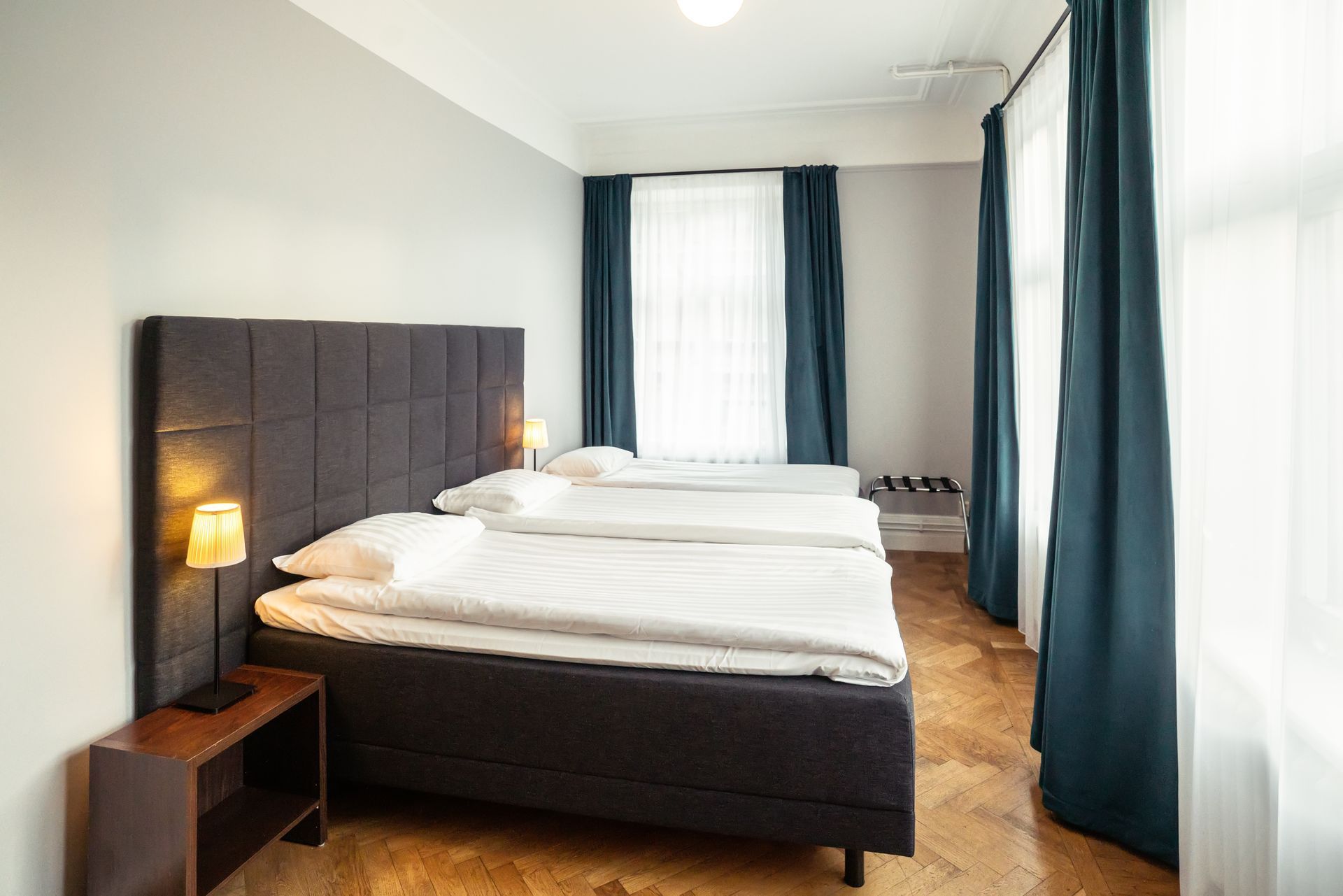 Bright bedroom with a dark upholstered bed, white bedding, and blue curtains by tall windows.