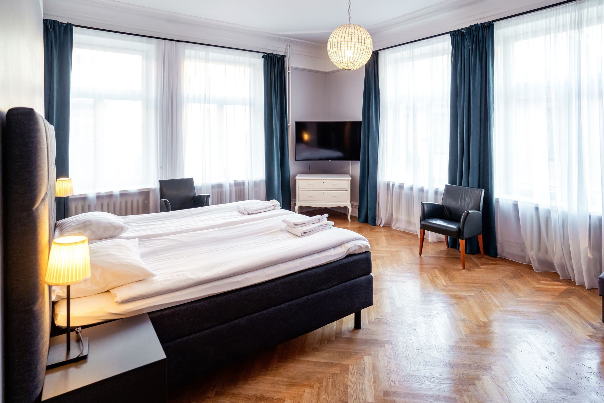 Bright modern bedroom with large windows, dark bed, wooden floor, and sitting area