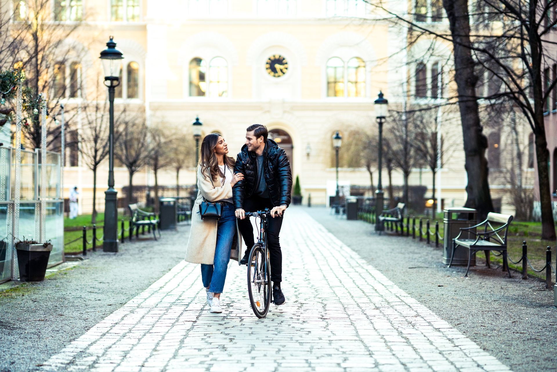 Couple walking a bicycle down a cobblestone path in a city park with benches and trees
