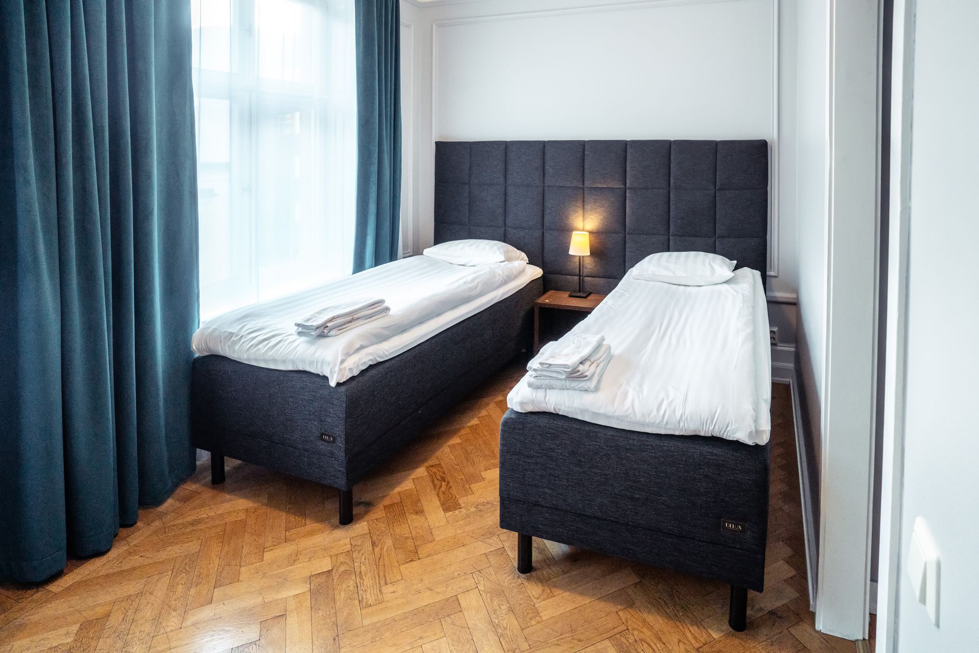 Two single beds in a bright bedroom with blue curtains and a lamp between dark upholstered headboards