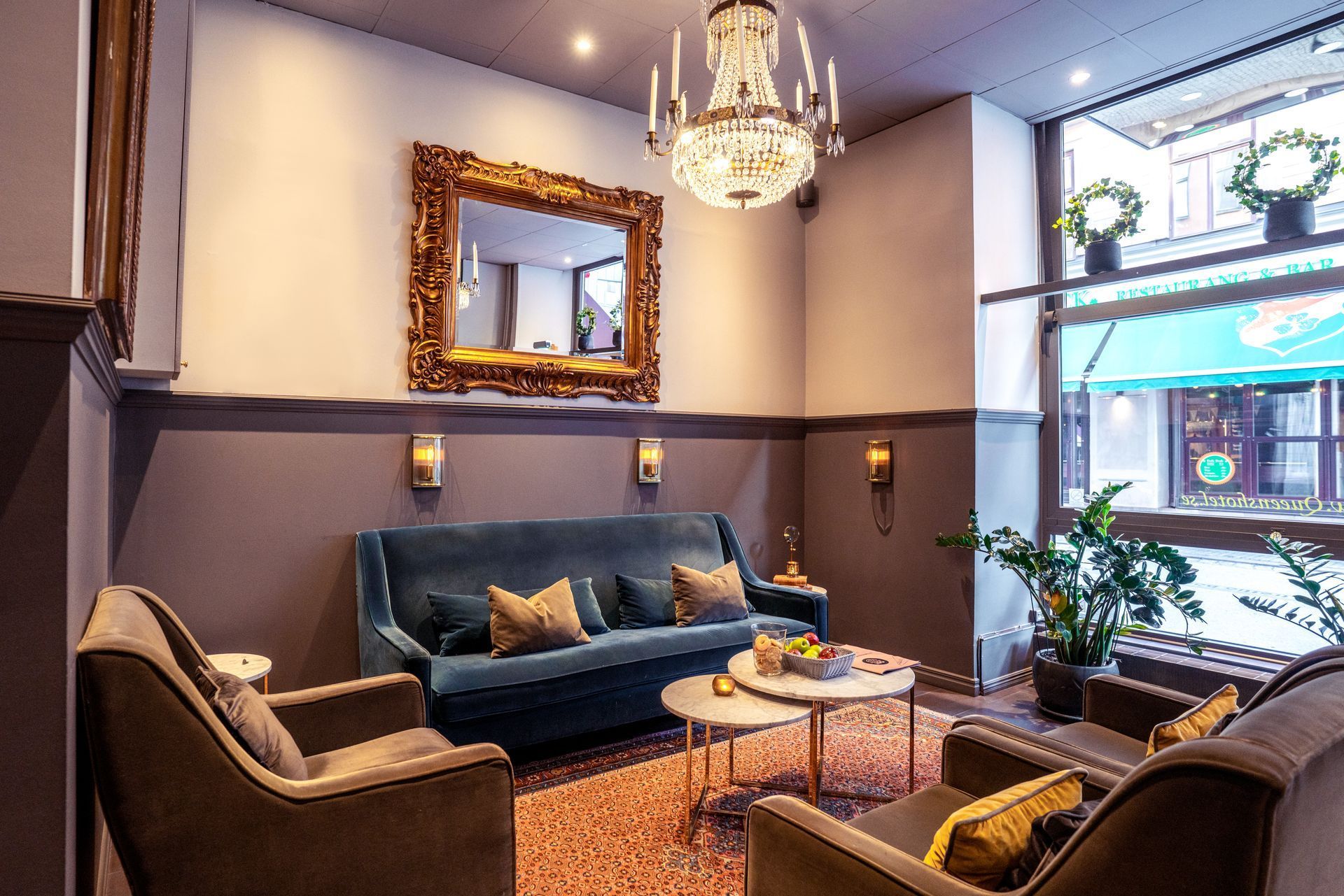 Cozy living room with dark sofa, armchairs, chandelier, large mirror, and window plants