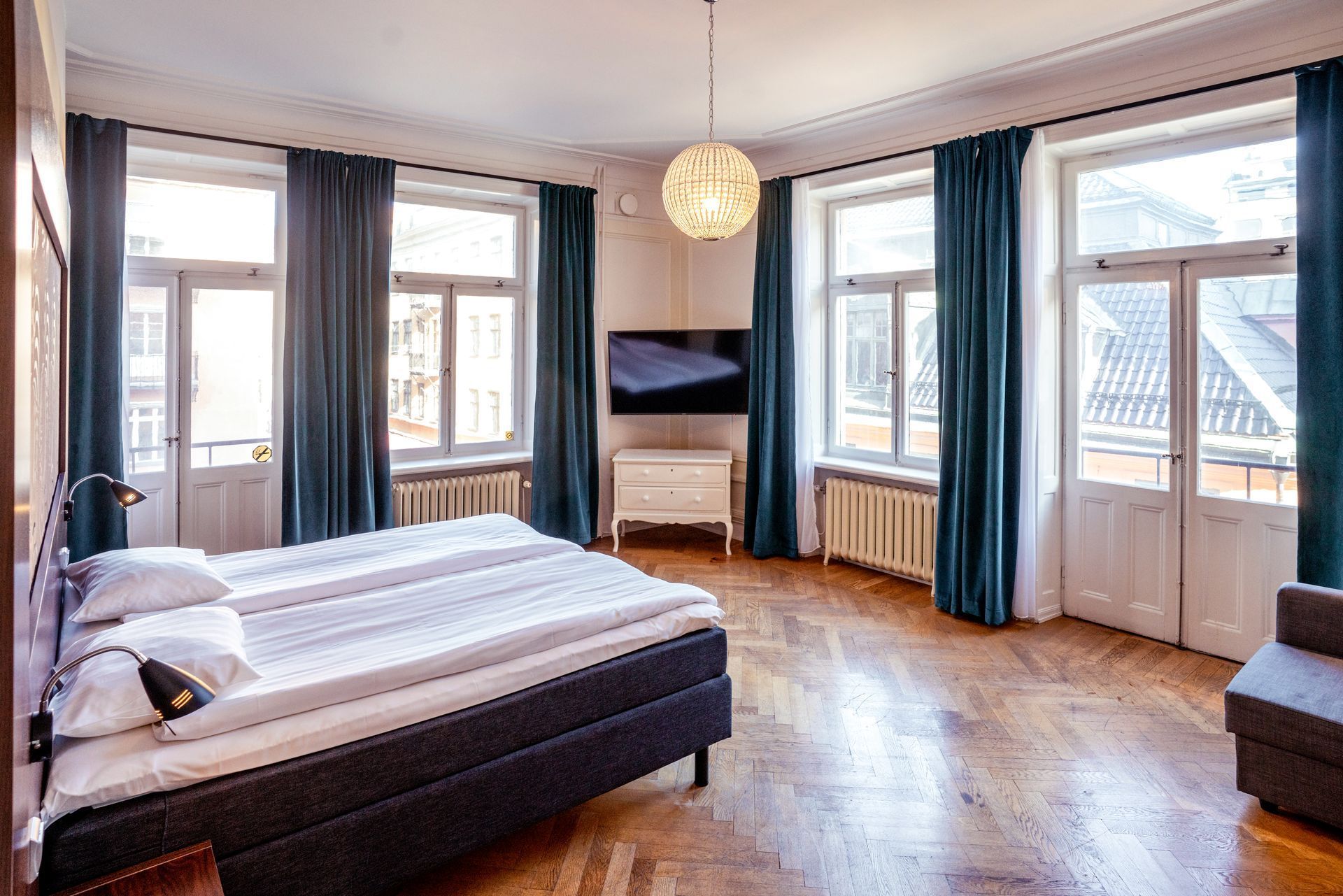 Bright bedroom with large windows, wooden floor, double bed, TV, and blue curtains