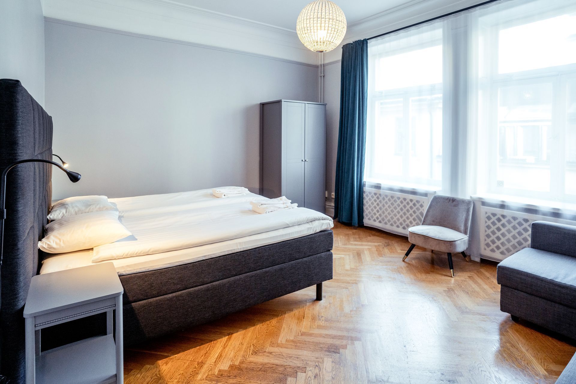 Bright modern bedroom with a bed, wardrobe, chair, hardwood floor, and large windows.