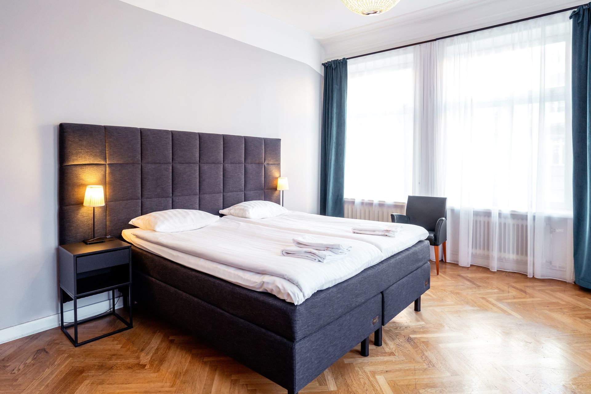 Bright bedroom with a dark upholstered bed, bedside lamps, and large windows with sheer curtains