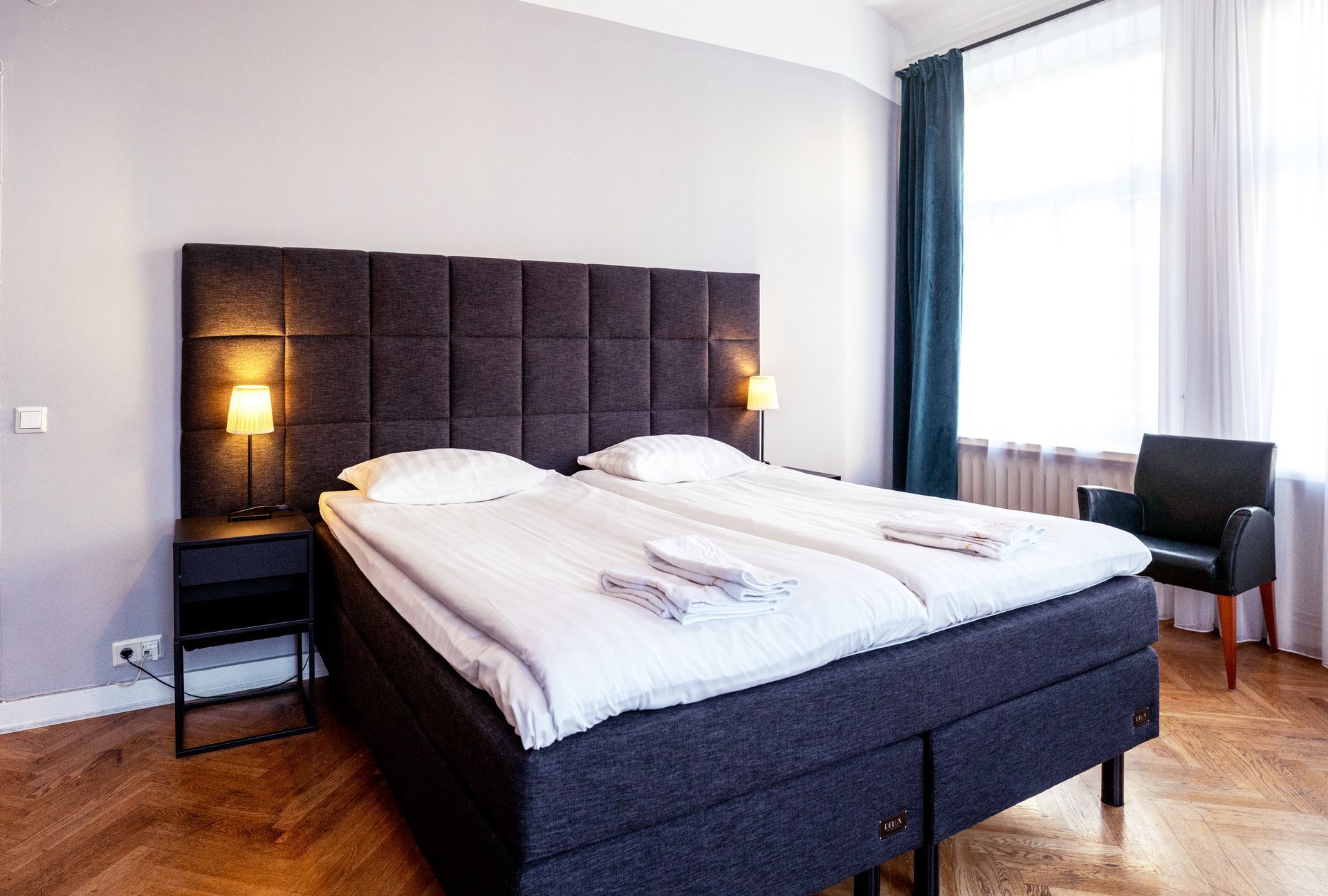 Modern bedroom with a large bed, dark upholstered headboard, bedside lamp, and bright window with curtains