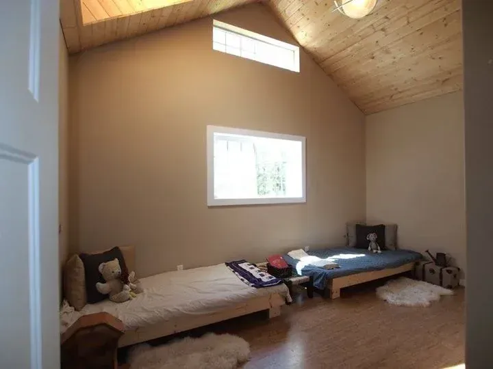 Bedroom with two low beds, tan walls, and a wood-plank ceiling. Two windows allow natural light to enter.