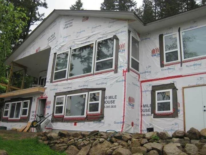 House under construction, windows installed, wrapped in white protective covering, stone wall in front.